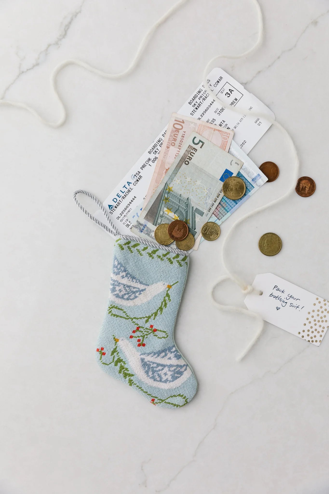 The Bauble Stockings Peace on Earth Needle Point Stocking is displayed on a white surface, filled with international banknotes, coins, airline boarding passes, and topped with a gift tag and string.
