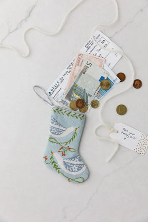 The Bauble Stockings Peace on Earth Needle Point Stocking is displayed on a white surface, filled with international banknotes, coins, airline boarding passes, and topped with a gift tag and string.