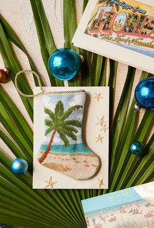 The Seaside Serenity Needle Point Stocking by Bauble Stockings features a hand-stitched palm tree design—perfect for adding coastal flair to your holiday decor. Displayed amid palm leaves, blue ornaments, postcards, and starfish.