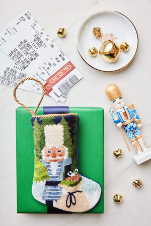 A green gift box with a Bauble Stockings Winter Solstice Nutcracker Needle Point Stocking, ballet tickets, gold jingle bells, and jewelry on a dish arranged on a white surface.