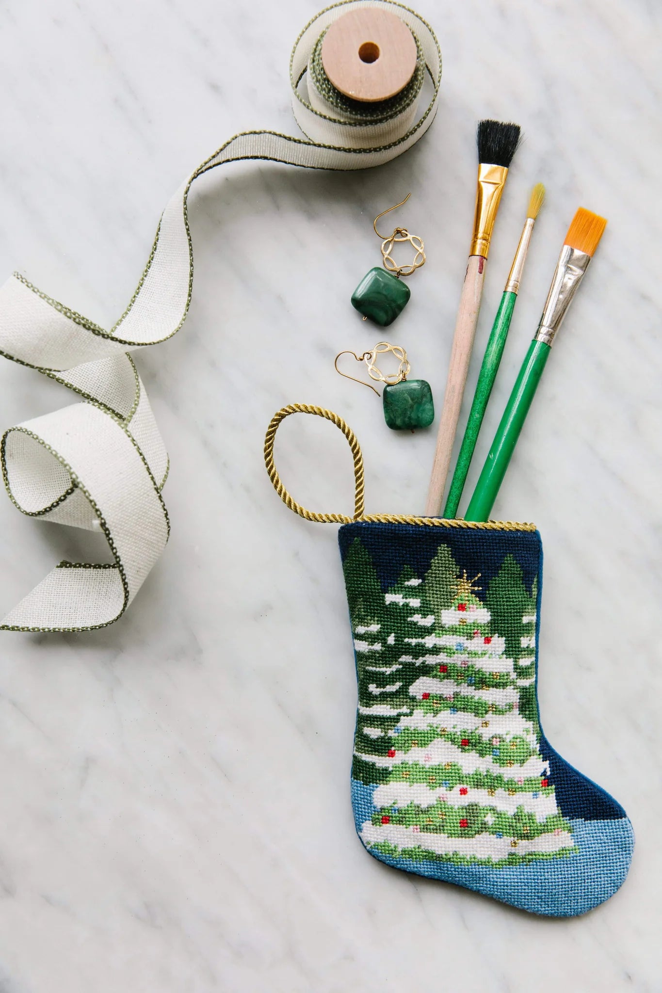 The Winter Wonderland Needle Point Stocking by Bauble Stockings, featuring a tree design, holds three paintbrushes and is displayed with ribbon and green stone earrings—a festive touch for your Christmas mantle.