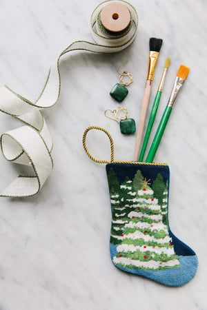 The Winter Wonderland Needle Point Stocking by Bauble Stockings, featuring a tree design, holds three paintbrushes and is displayed with ribbon and green stone earrings—a festive touch for your Christmas mantle.