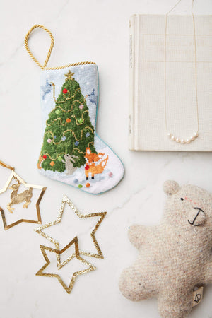 The Woodland Creatures Needle Point Stocking by Bauble Stockings, paired with gold star ornaments, a plush bear, a pearl necklace, and a closed book, creates a charming and festive holiday display on any white surface.