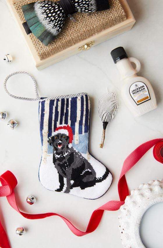 The Woodland Frolic Needle Point Stocking by Bauble Stockings features a hand-stitched black dog in a Santa hat surrounded by festive ribbons, a bottle, small bells, and decorative accents on a white background.