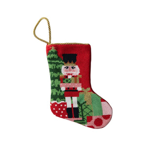 The Classic Nutcracker Needle Point Stocking by Bauble Stockings is a red hand-stitched stocking featuring a nutcracker, Christmas tree, and presents, finished with a gold hanging loop—ideal as a festive ornament or gift.