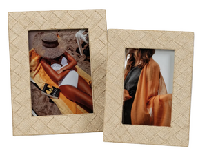 Two woven raffia frames from the Zodax La Bocca Frame Collection add natural chic charm—one displays a sunbathing woman on the beach, the other features a woman indoors with an orange shawl.
