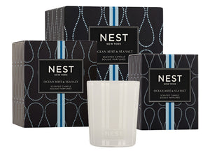 A white Nest Ocean Mist & Sea Salt Candle from the Ocean Mist & Sea Salt Candle Collection is displayed in front of two black patterned boxes, evoking the refreshing aroma of ocean mist and sea salt.