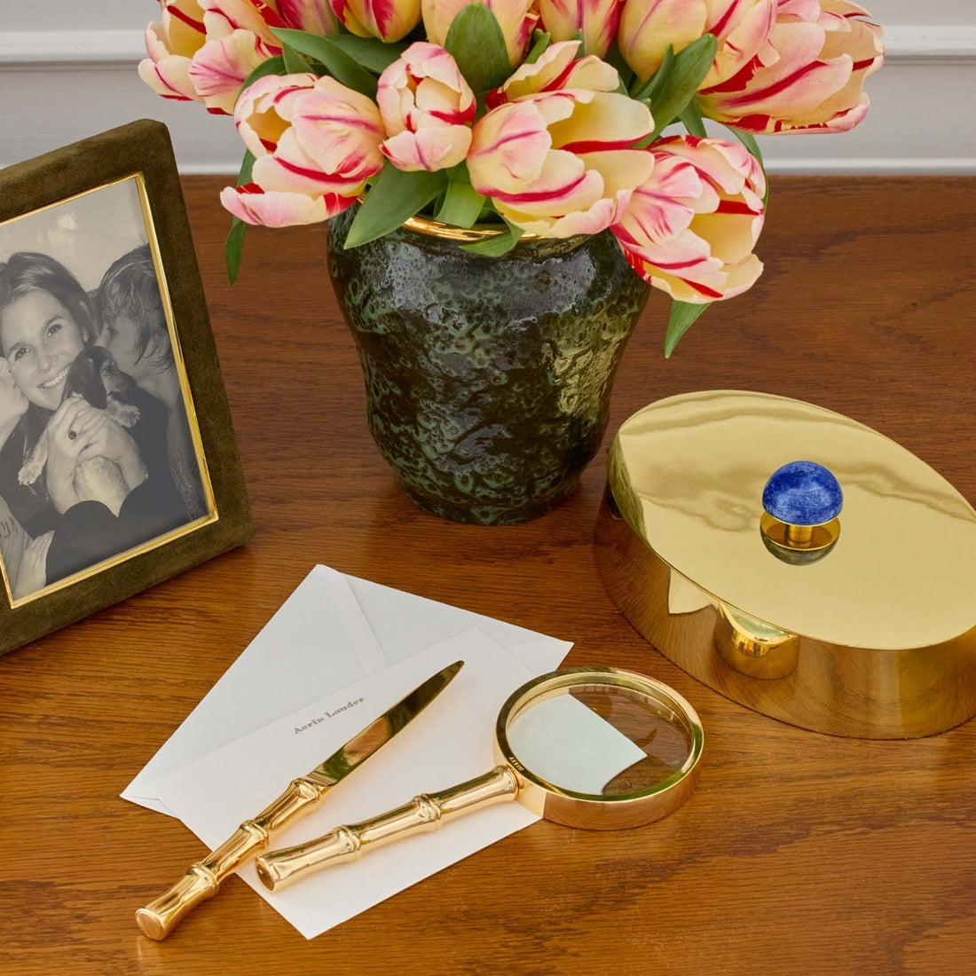 On a wooden desk sits Aerin's Ava Bamboo Magnifying Glass and Letter Opener in gold, a framed black-and-white photo, a vase of pink tulips, a gold box, and two white envelopes.