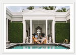 A group of people sit around an outdoor table with drinks at the Nick Mele Fine Art Poker by Nick Mele Photography location, surrounded by classical white columns and neatly trimmed green hedges, next to a rectangular pool with blue water in the foreground.