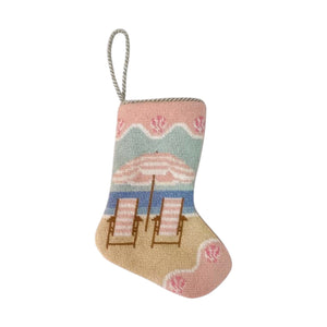 The Bauble Stockings Palm Beach Holiday Needle Point Stocking features a beach scene with two lounge chairs and a pink-striped umbrella. This small ornament comes with a hanging loop, making it a perfect Christmas gift or family tradition.