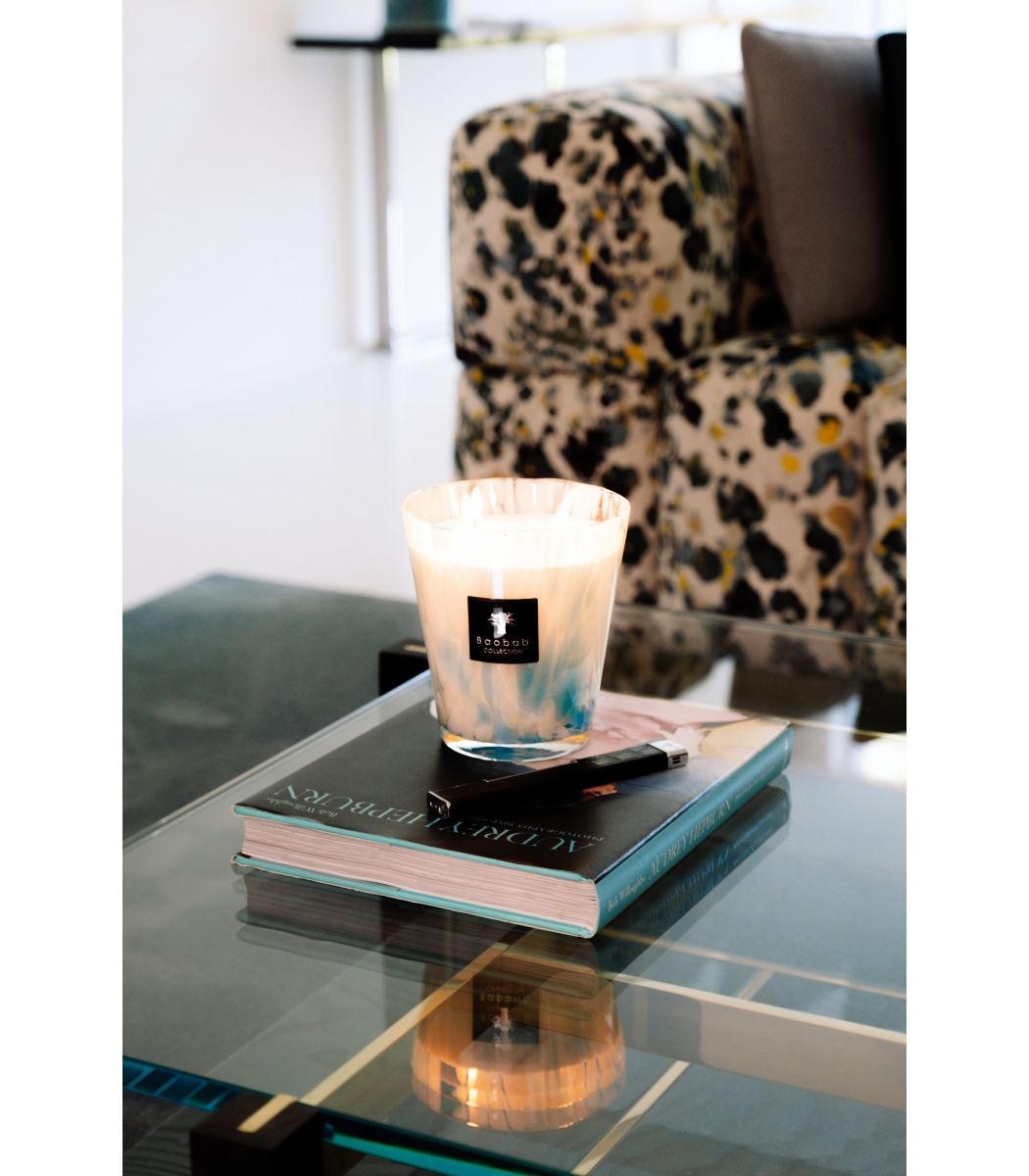 A Baobab Collection Pearls Sapphire Candle in a glass holder sits atop a coffee table book on a reflective glass table, with a patterned chair in the background.