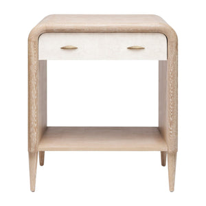 The Pierre Nightstand Collection by Made Goods features a whitewashed oak piece with a rounded frame, tapered legs, an open lower shelf, and a single white drawer accented by two brass handles.