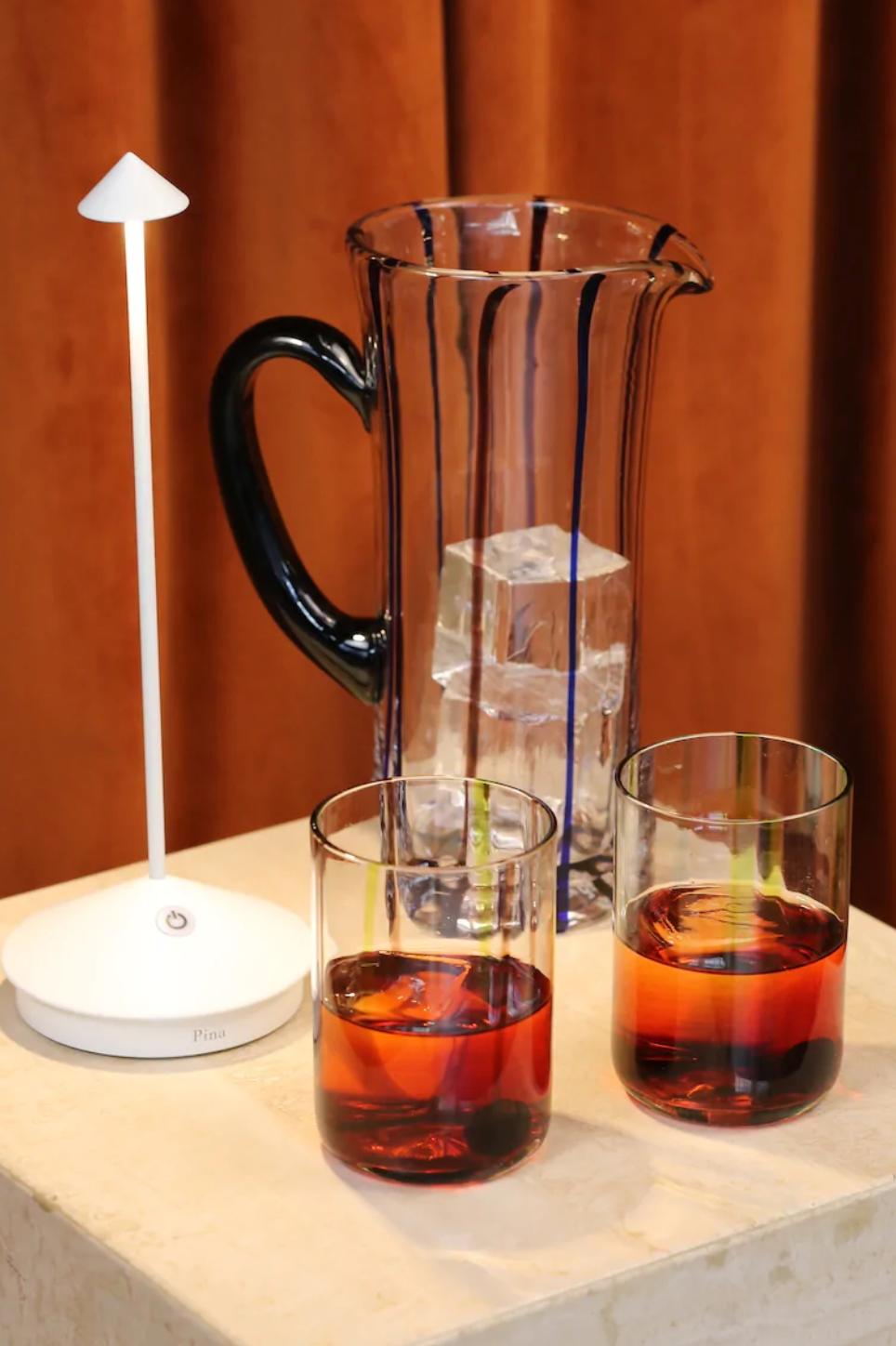 The Zafferano America Pina Lamp Collection touch-dimmable portable rechargeable lamp sits on a beige surface by a glass pitcher and two amber drinks, set before a brown curtain—perfect for indoor or outdoor use.