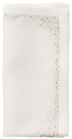 The Kim Seybert Pin Dot Napkin Set features a folded white napkin with a decorative border of small gold and silver dots along two adjacent edges, offering an elegant design.