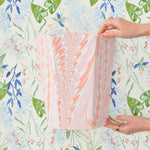 Two hands hold a pastel Marble Bath Collection item by Weezie Towels, crafted from recycled paper, in front of floral-patterned wallpaper with green leaves and blue flowers.