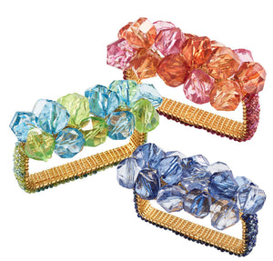 Three rectangular Kim Seybert Pop Rocks Napkin Rings feature gold bands with clusters of large faceted beads in pink-orange, blue-green, and light blue-purple, creating a sparkling, bejeweled design.