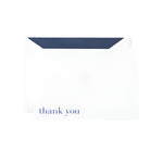 The Crane Bodoni Thank You Folded Note Card Set features white thank you cards with Bodoni Blue text, partially inserted into a dark-lined envelope on a plain background. The use of thermography gives the Thank You Note a textured and classic finish.