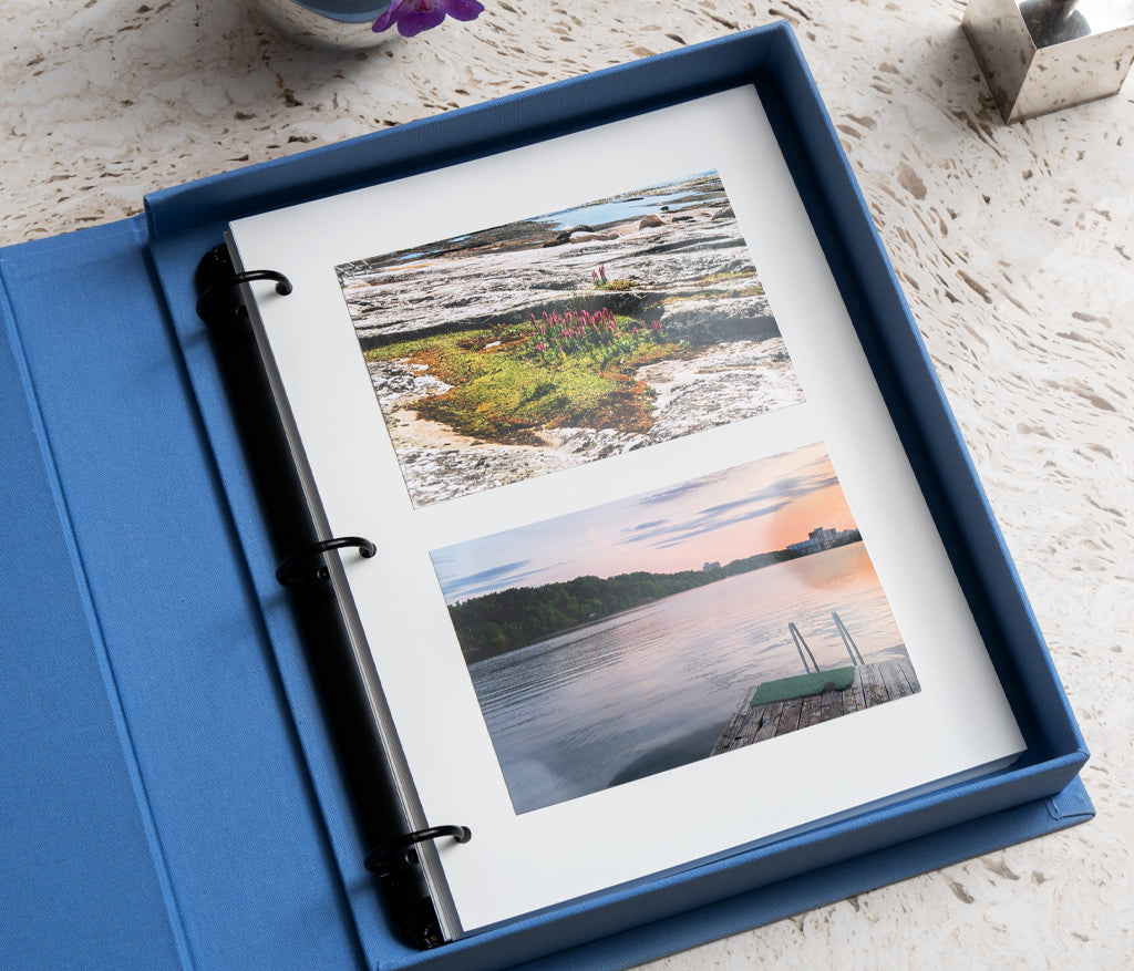 The PrintWorks Moments That Matter The Most Photo Album features an open blue cotton cover coffee table design displaying two landscape photos—rocky terrain with vegetation and a sunset lake dock—on high-quality photo paper.