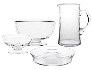 The Juliska Puro Glass Serveware Collection features a clear pitcher, large bowl, small dessert dish, and round plate with raised center—each mouth-blown with elegant textural lines and showcased on a white background.