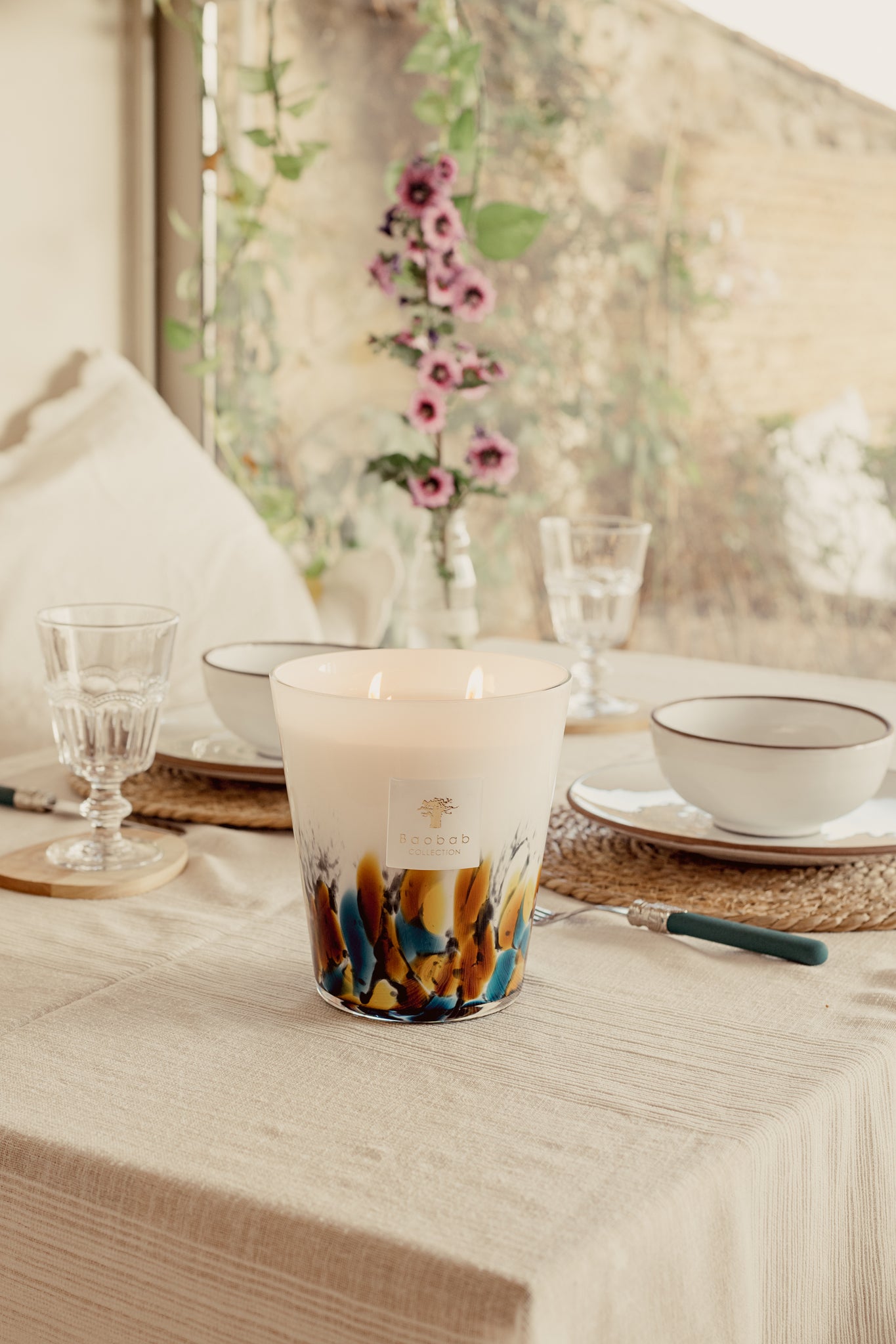 A large Baobab Rainforest Mayumbe scented candle on a table set with dishes and glasses, near a floral arrangement, in a warm, well-lit room.