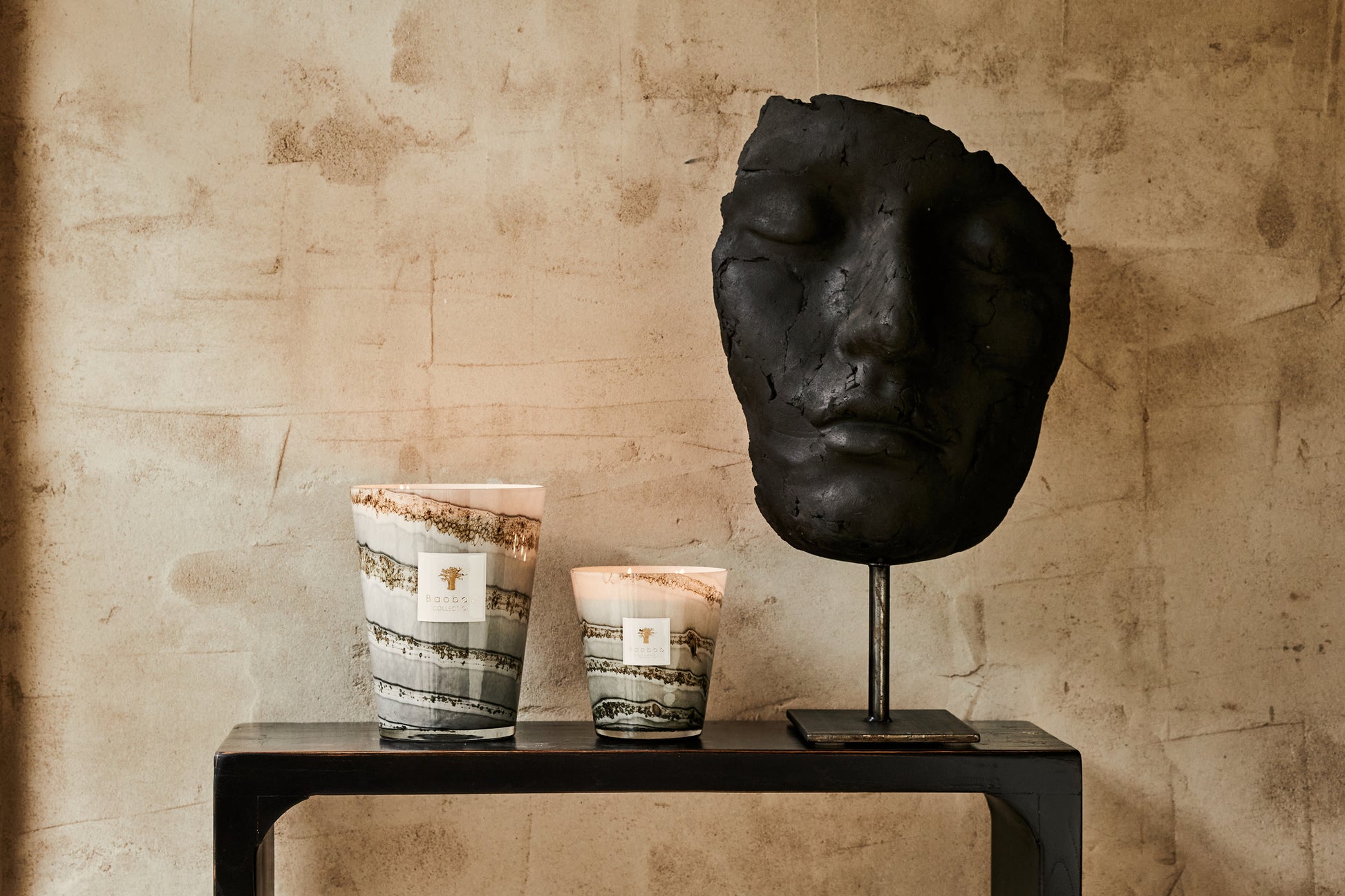 A large black sculpted head on a stand beside two Baobab Collection Sand Atacama candles on a narrow table against a textured beige wall.