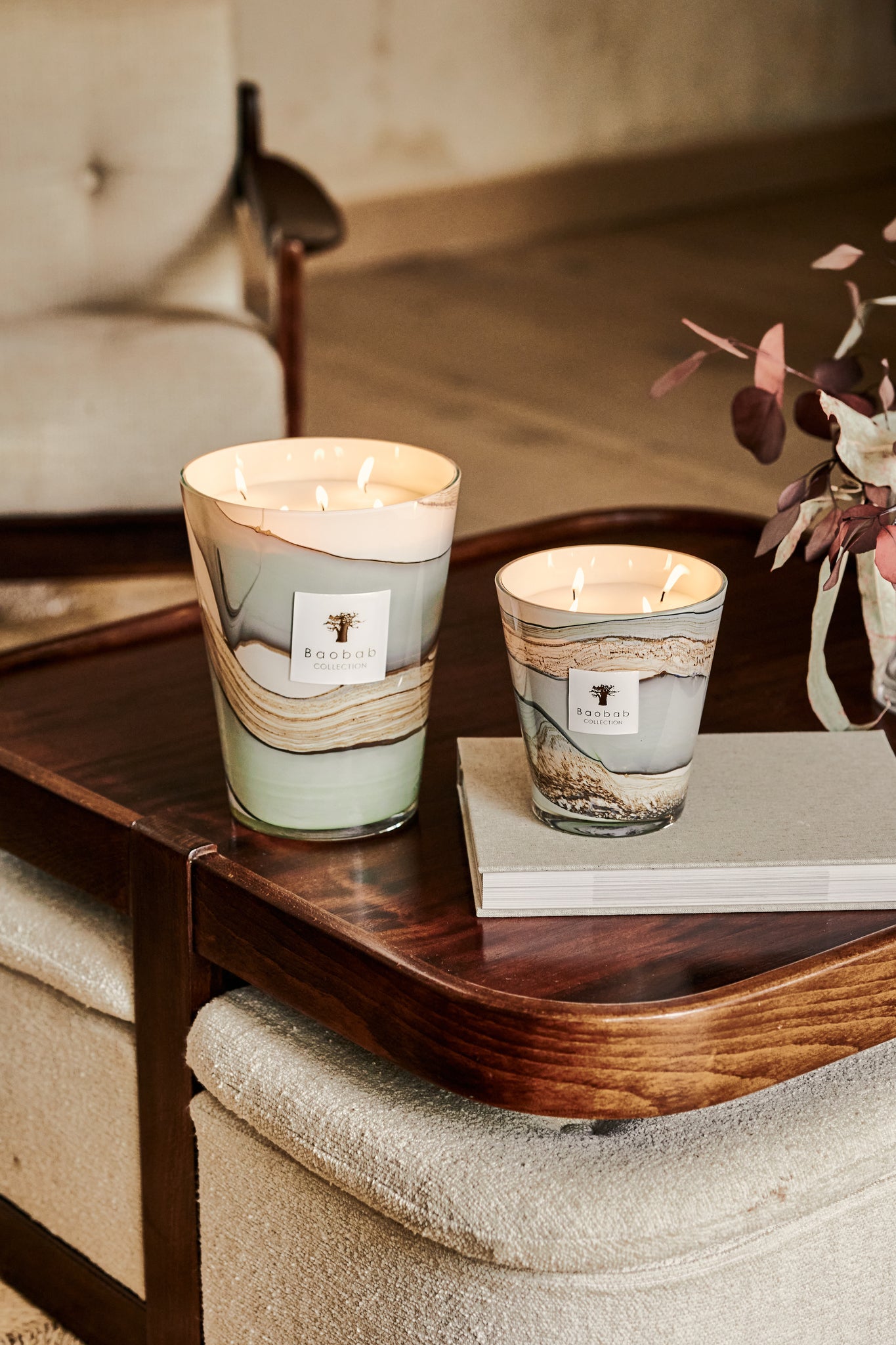 Two Baobab Collection Sand Sonora candles on a wooden coffee table, alongside a book and floral decoration, in a cozy indoor setting.