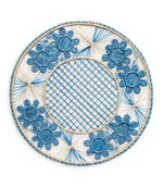 Handcrafted in Colombia, the Serenity Rattan Placemat Collection by Themis Z features round placemats with blue and beige woven patterns, blue floral designs along the rim, and a crisscross center pattern.