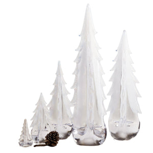 The Simon Pearce Snowy Branches Evergreen Tree Collection features five clear glass trees of varying heights arranged in a row, with a small pine cone in front, evoking the charm of Vermont’s winter woods.