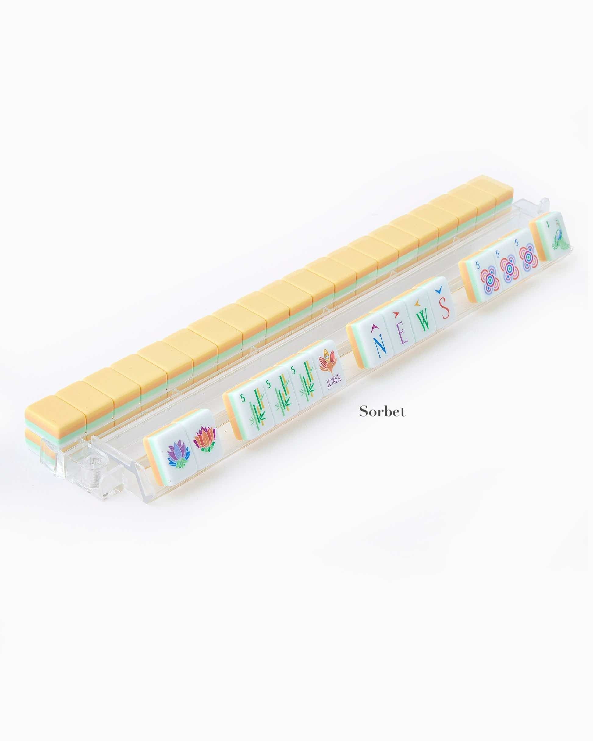 A clear rack holds yellow-topped, hand-painted tiles from the Oh My Mahjong Mahjong Tile Collection, with seven tiles in front showing assorted designs and the word "NEWS." The set is labeled "Sorbet.