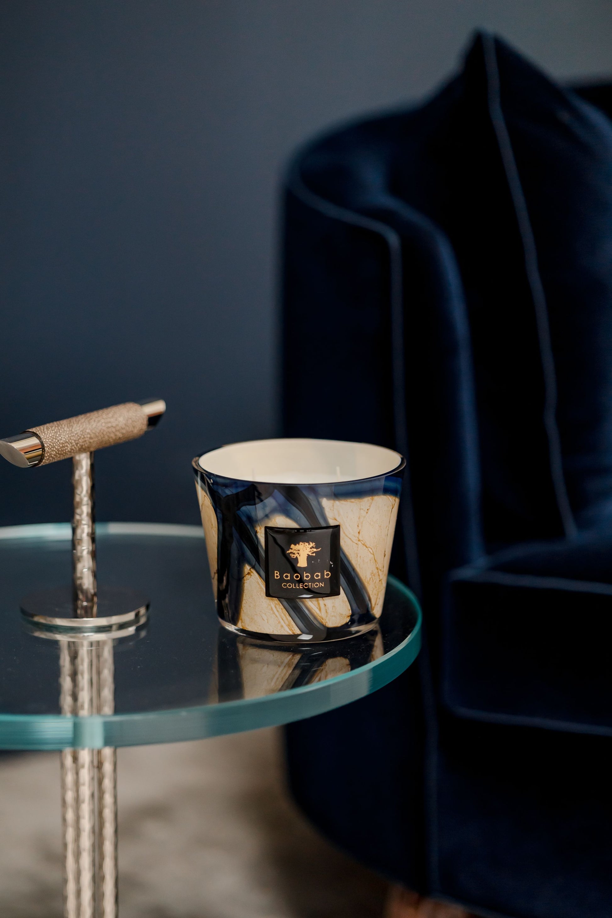 A Baobab Collection Stones Lazuli Candle by Baobab with a dark marbled container sits on a glass side table with a metallic base, next to a navy blue velvet chair.
