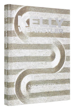 The image shows the cover of the Rizzoli book "Kelly Wearstler: Synchronicity," featuring an abstract design with parallel lines and curves in neutral tones. The title and author's name are displayed in large metallic text, reflecting her renowned expertise in both residential and hospitality designs.