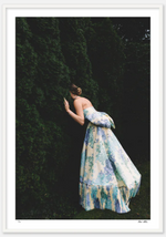 A woman in a flowing floral gown from "Nick Mele Fine Art Alice" by Nick Mele Photography peers into a dense hedge, partially hiding her face, amidst the lush backdrop of Palm Beach.