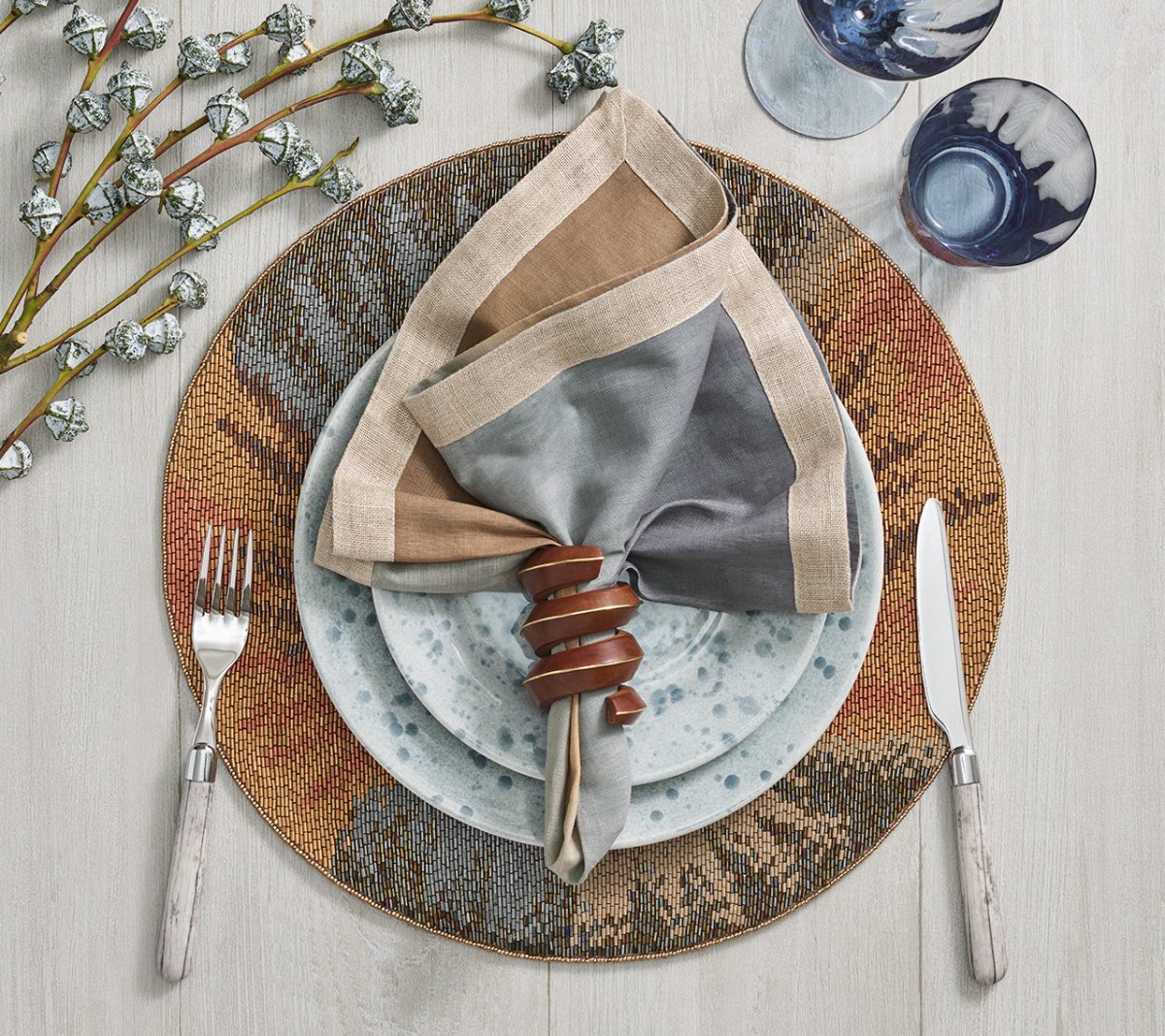 A place setting with a fork, knife, patterned plates, two blue glasses, a multicolored napkin in the hand-carved Kim Seybert Tendril Napkin Ring, and decorative branches on a light wood table.