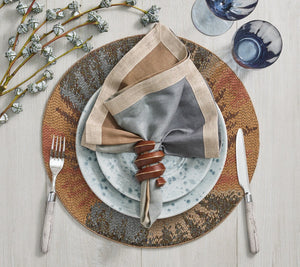 A place setting with a fork, knife, patterned plates, two blue glasses, a multicolored napkin in the hand-carved Kim Seybert Tendril Napkin Ring, and decorative branches on a light wood table.