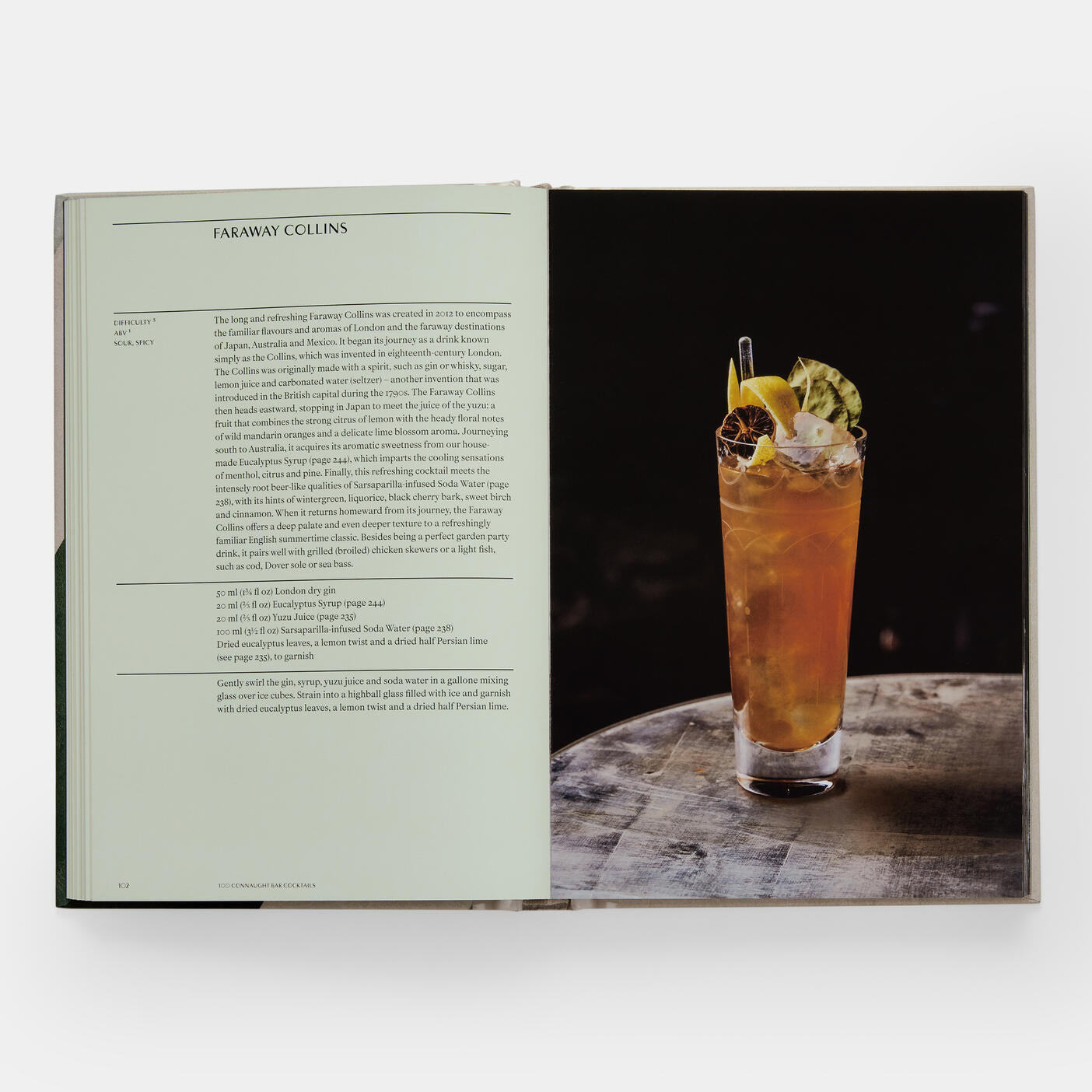 An open "The Connaught Bar: Recipes and Iconic Creations" by Phaidon shows a Faraway Collins recipe crafted by expert mixologists on the left, with a tall glass of the cocktail garnished and resting on a wooden surface, echoing renowned creations from places like Connaught Bar.
