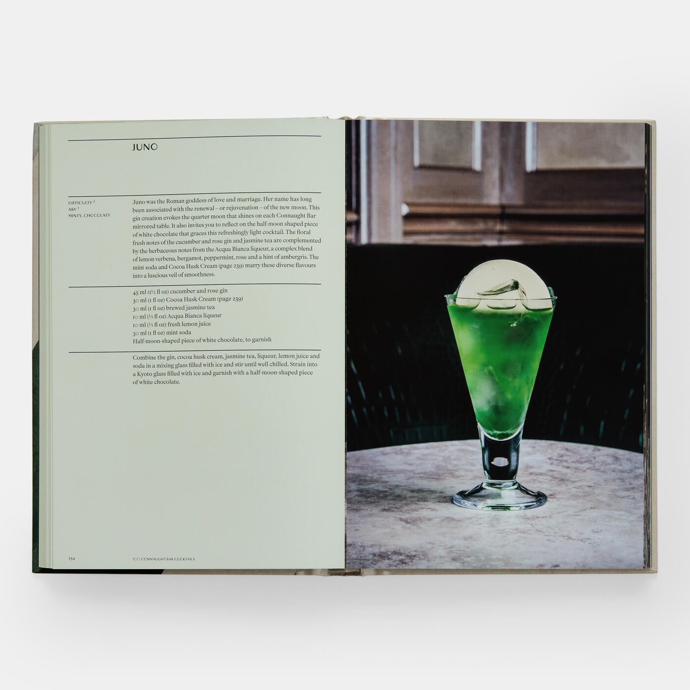 A Connaught Bar cocktail recipe from Phaidon's "The Connaught Bar: Recipes and Iconic Creations" lies open to the left, with a vivid green drink artfully prepared by expert mixologists showcased in a glass on the right, both elegantly set on a marble table.