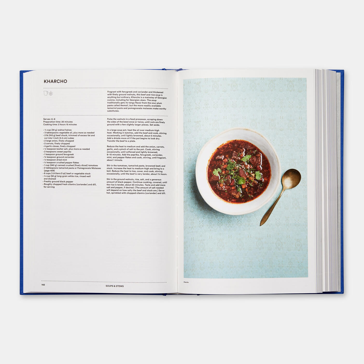 Open "The Jewish Cookbook" by Phaidon displaying a kharcho soup recipe on the left and an image of the soup garnished with herbs in a white bowl on the right, with a gold spoon beside it—an inviting blend of home-cooking recipes and international tastes.