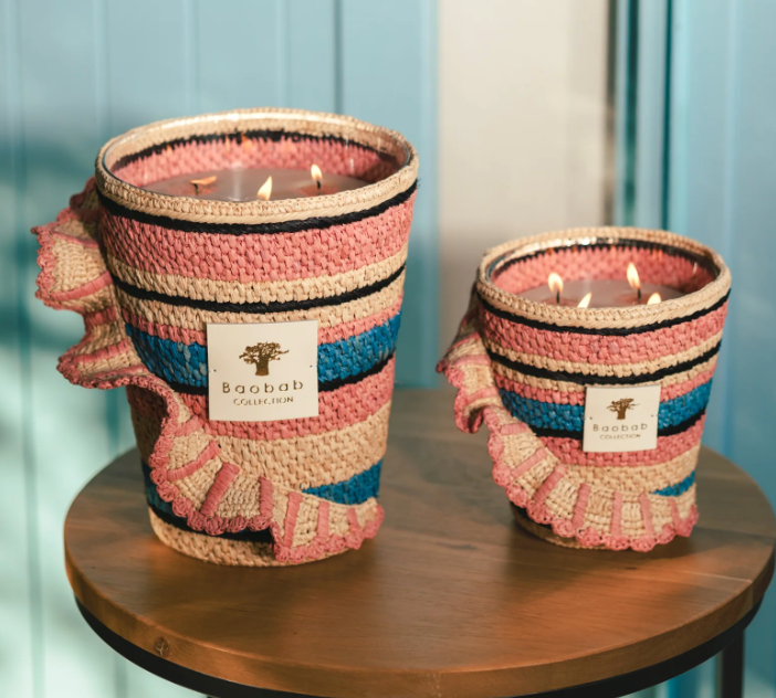 Two Baobab Collection Fety Mandihy scented candles in pink, blue, and beige woven holders sit on a wooden table. Each has multiple wicks and a rectangular label against a light blue background, enhancing their charm.