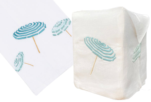 Two images of a white Italian linen napkin from the Beach Umbrella Collection, Aqua by Haute Home hand embroidered with teal and blue beach umbrellas; one image shows the napkin flat, the other folded upright.