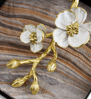 The Michael Aram Orchid Cocktail Napkin Holder features a gold and white metal floral design with two blossoms and three buds, resting on brown, beige, and gray striped fabric—a touch of subtle luxury and elegance.