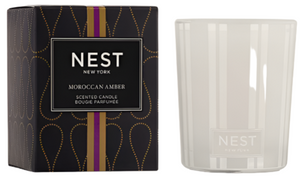 A box labeled "Moroccan Amber Candle Collection" by Nest sits beside a white glass candle holder with the Nest logo, evoking rich patchouli and bergamot notes in its elegant design.