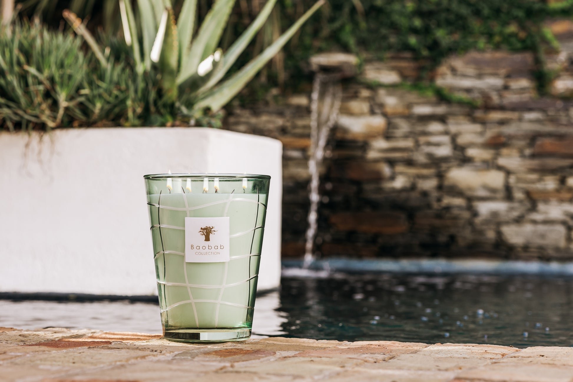 A large green Baobab Collection Waves Nazare Candle on a stone patio with a background water feature and lemon tree wood plants.
