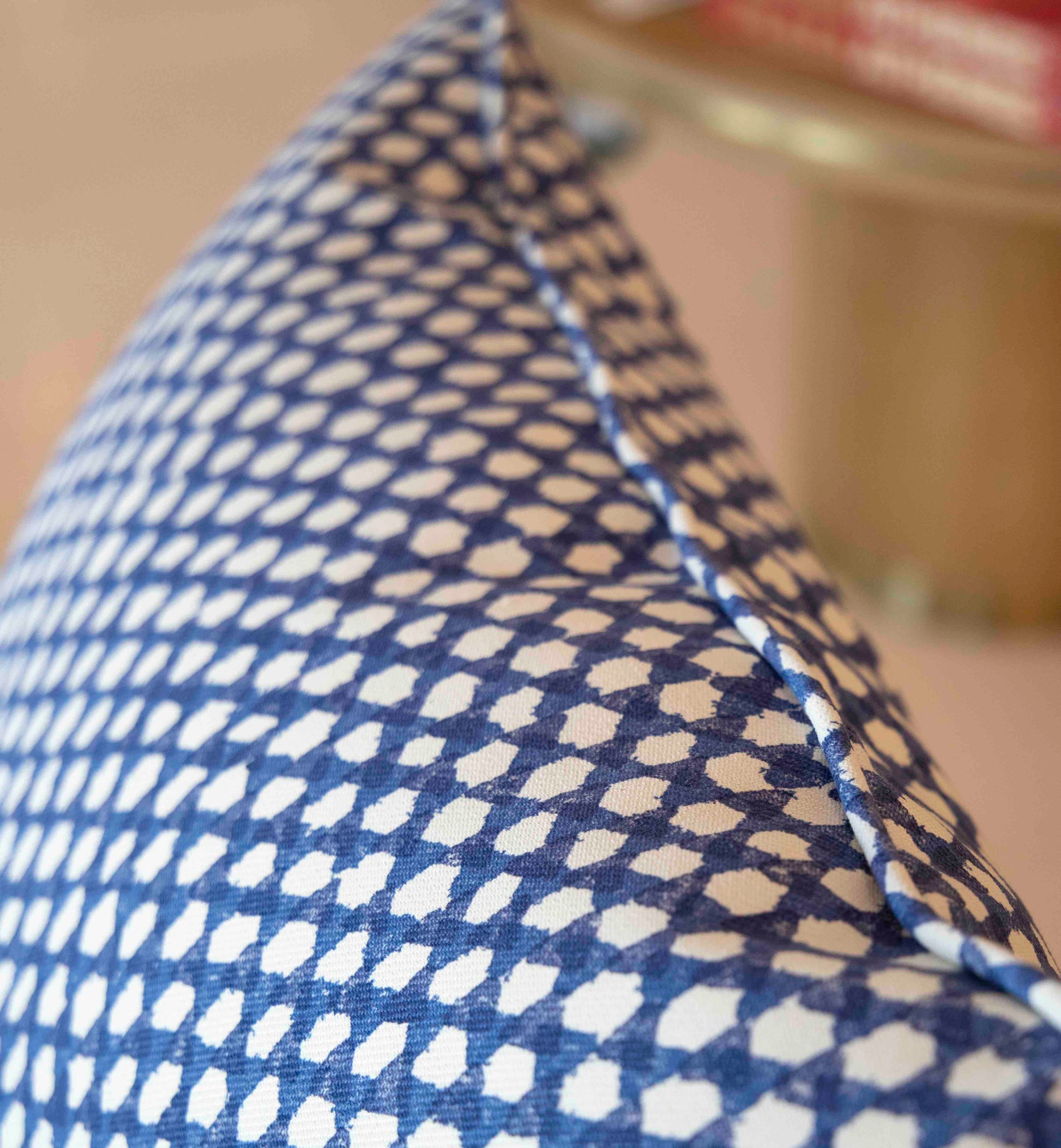 Close-up of the Royal Blue Wicker Outdoor Pillow by Associated Design features a blue and white pattern with visible edge stitching; a blurred table with books appears in the background.