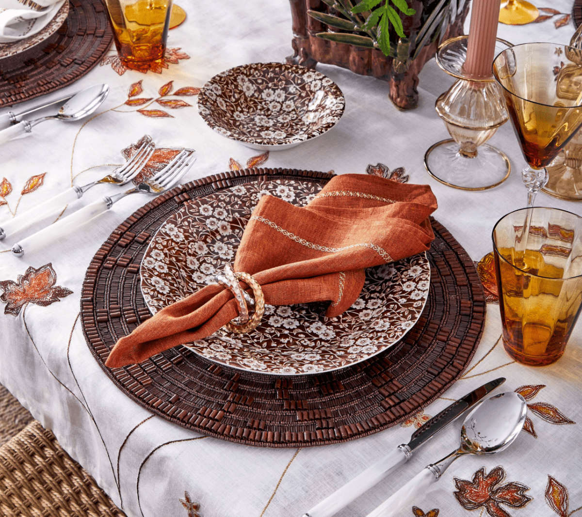 Elegant autumn table setting featuring Kim Seybert's Wood Matrix Placemat, brown floral plates, an orange napkin with a ring, amber glasses, silver cutlery, and a white tablecloth with leaf embroidery.