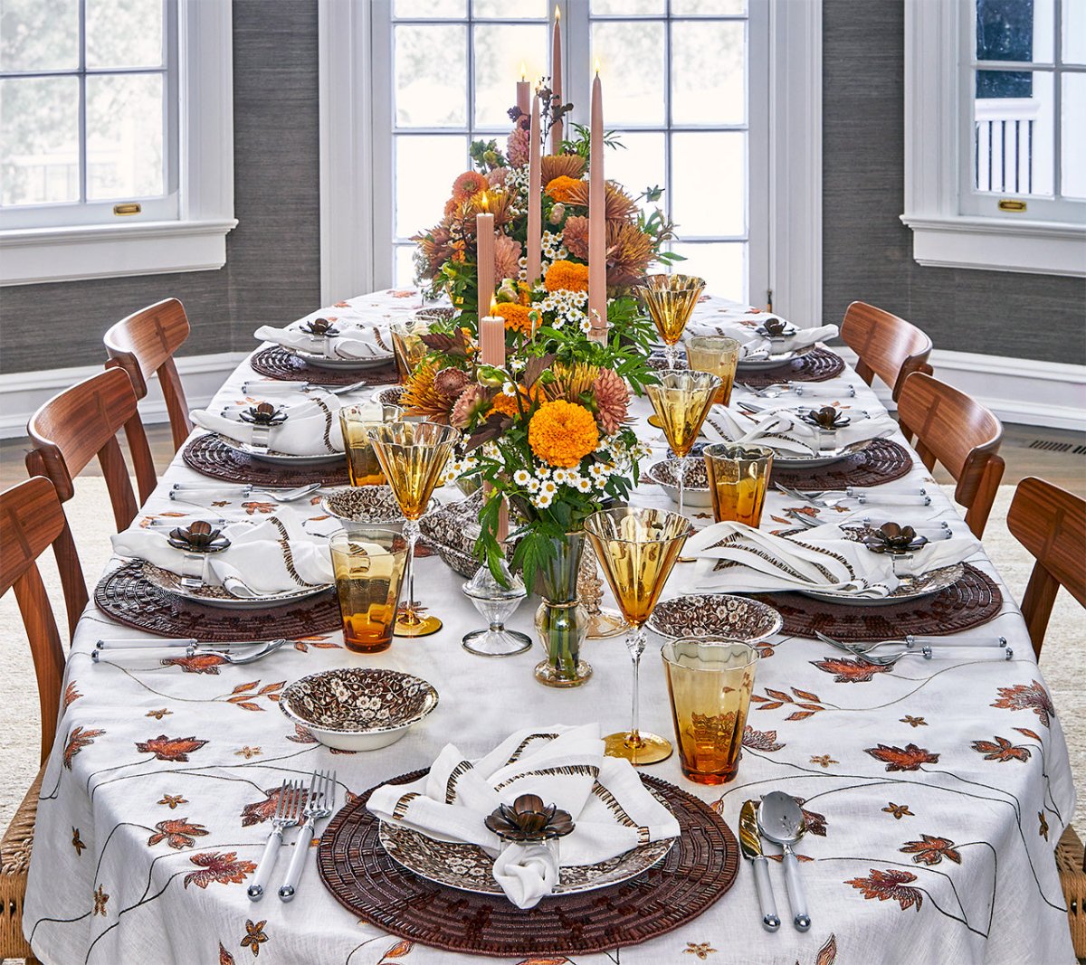 Formal dining table set for a meal with autumn décor, floral centerpieces, orange and yellow candles, glassware on a leaf-patterned cloth, and Kim Seybert Wood Matrix Placemats accented with handcrafted wooden beads.