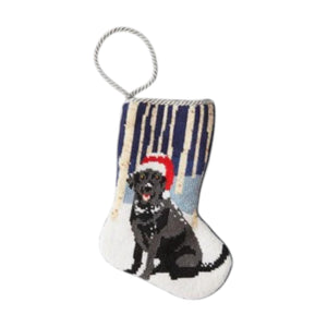 The Woodland Frolic Needle Point Stocking by Bauble Stockings features a hand-stitched black dog in a red Santa hat, sitting amid snowy trees, perfect for adding festive charm to your Christmas décor.