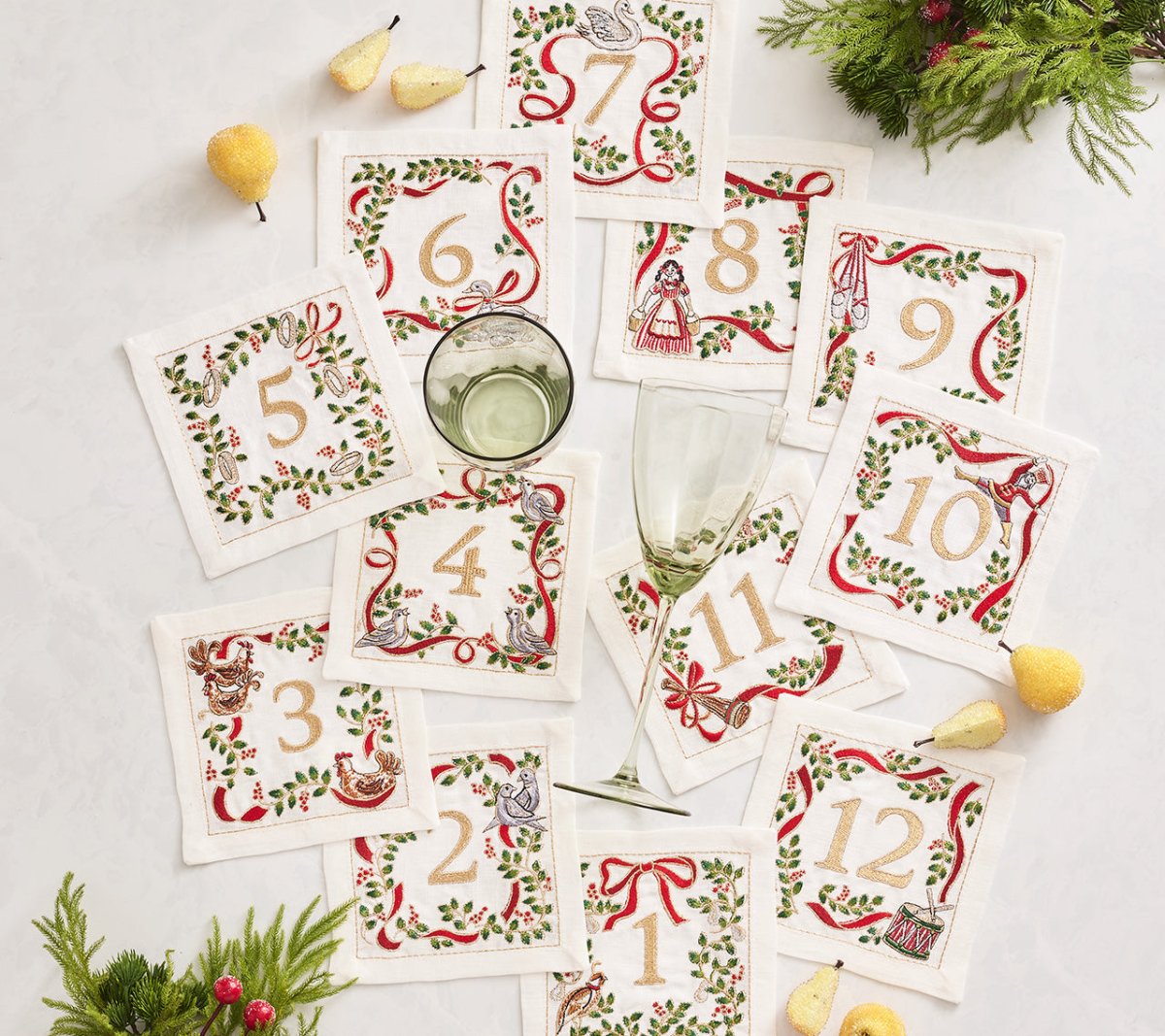 The Kim Seybert Christmas Carol Cocktail Napkins Set, numbered 1 to 12 with embroidered borders, is beautifully styled with wine glasses, yellow fruit, and festive greenery—ideal for holiday gatherings on a white surface.