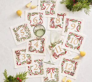 The Kim Seybert Christmas Carol Cocktail Napkins Set, numbered 1 to 12 with embroidered borders, is beautifully styled with wine glasses, yellow fruit, and festive greenery—ideal for holiday gatherings on a white surface.