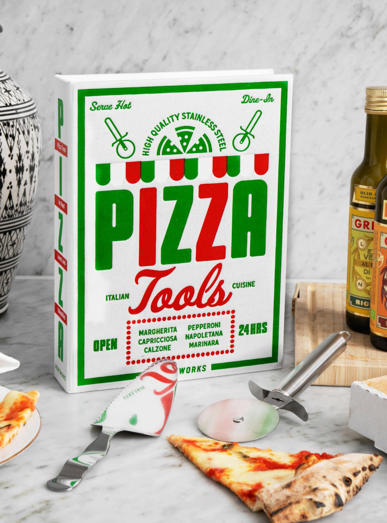 The Essentials Pizza Tools by PrintWorks, crafted from stainless steel and packaged in a pizza box design, sit on a marble counter with pizza slices and accessories—an essential upgrade to any pizza kit.