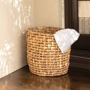 The Astoria Natural Round Basket by Pigeon & Poodle, 15" wide and crafted from water hyacinth, sits on a wooden floor near a wall and window with a white towel draped over its edge.